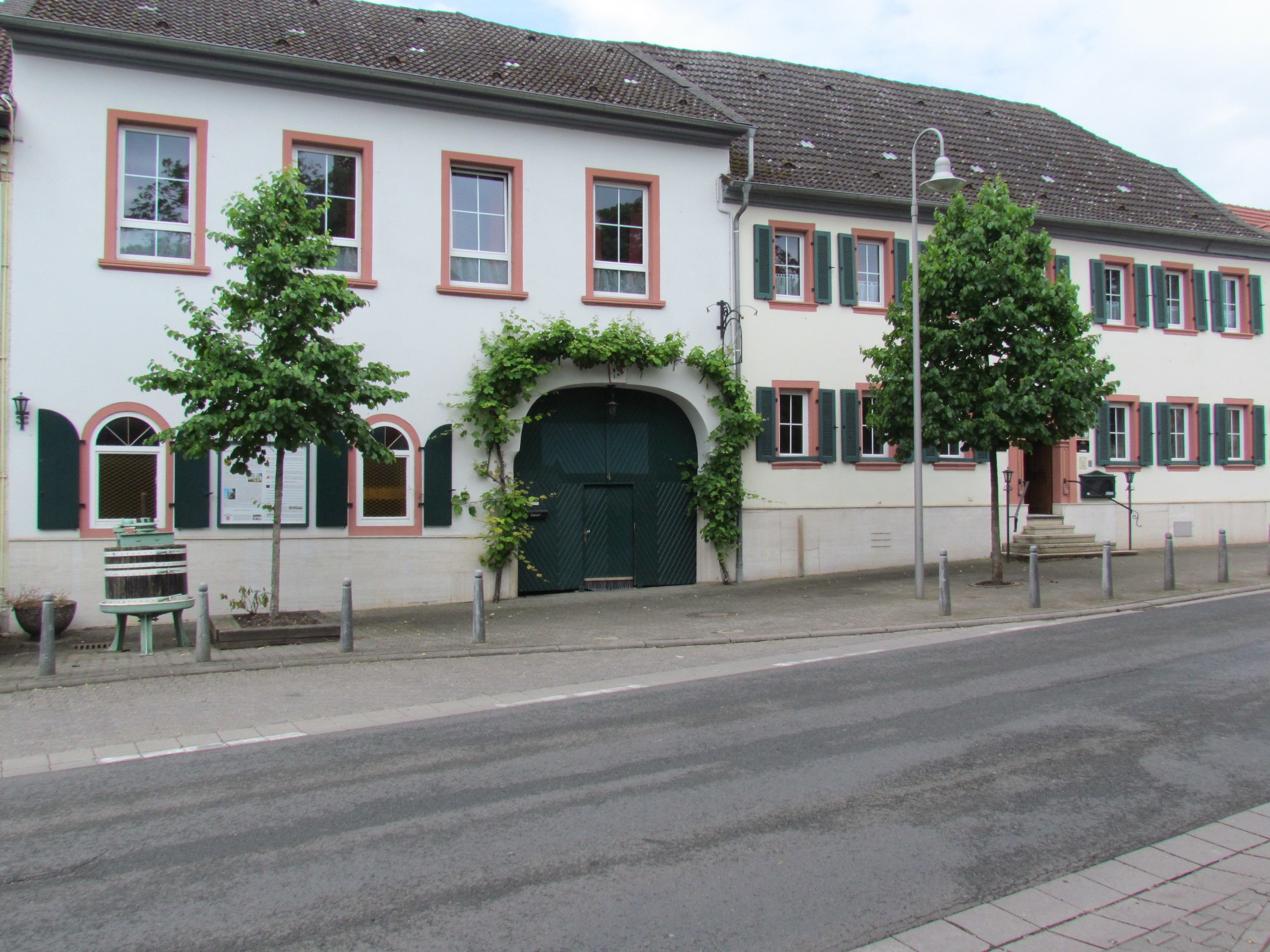 Ein zweistöckiges Gebäude mit einer grünen Tür, weißen Wänden und rotem Zierstreifen. Vor dem Gebäude befinden sich Bäume und eine Straße.