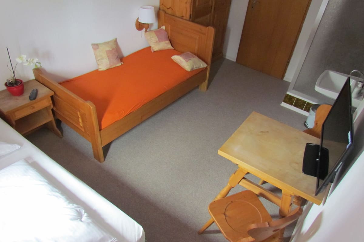 Une petite chambre avec un lit simple, un bureau avec une chaise, une télévision et une table de chevet avec une lampe.
