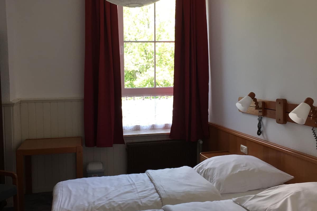 Une chambre d’hôtel avec deux lits, une tête de lit en bois, une table de chevet, une fenêtre avec des rideaux rouges et une lampe suspendue.