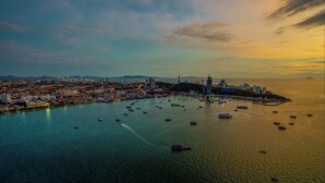 A coastal cityscape with a marina filled with boats, a prominent building with a blue sign, and a distant mountain range. - Joe Palace Beach Living Jomtien Pattaya (Pattaya)