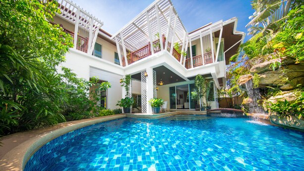 A swimming pool with a tiled floor, surrounded by lush greenery and a white building with balconies. - PARADISE Pool Villa Pattaya in Tropicana Village (Pattaya)