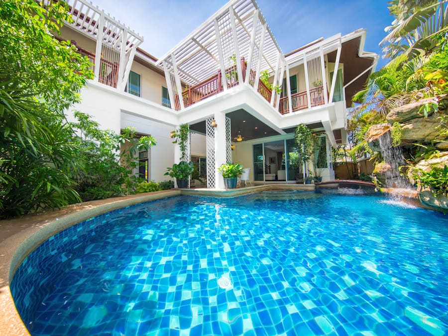 Una piscina con suelo de baldosas, rodeada de abundante vegetación y un edificio blanco con balcones.