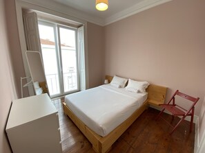 Une chambre à coucher avec un lit en bois, une table de chevet blanche, une chaise rouge et une grande fenêtre donnant sur des immeubles.