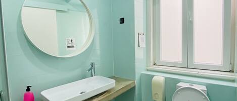 Une salle de bain avec un lavabo blanc, un miroir rond, des toilettes et une fenêtre.