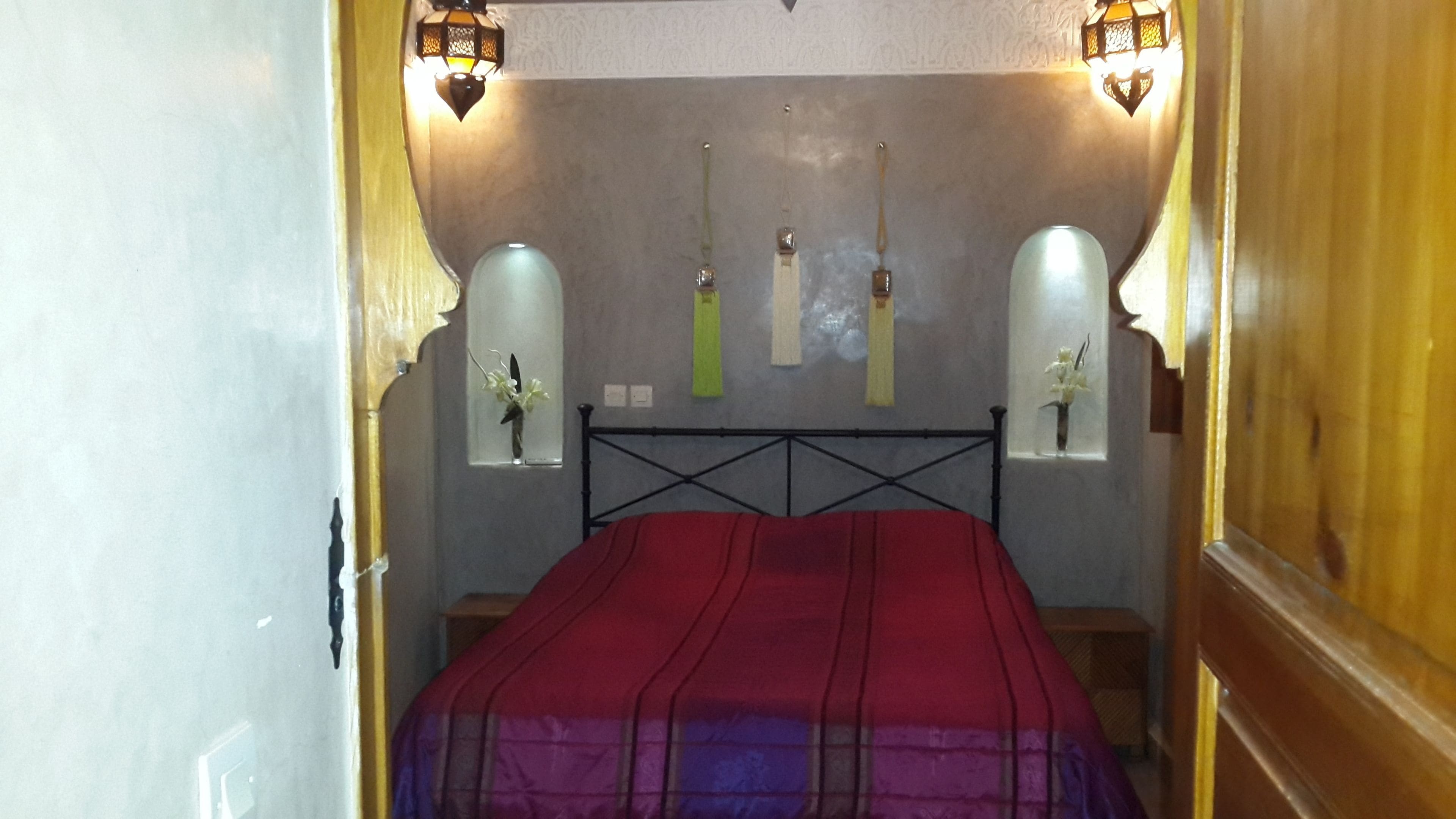 A narrow room with a bed, two wall-mounted lamps, and a small window with a vase of flowers.