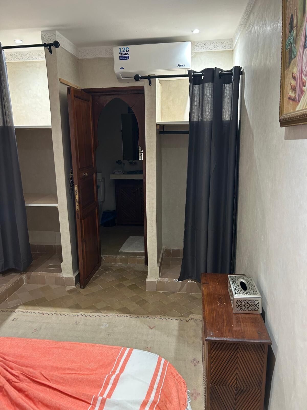 Un dormitori amb un llit, una nitrera de fusta, aire condicionat fixat a la paret i una porta que dona a una altra habitació.
