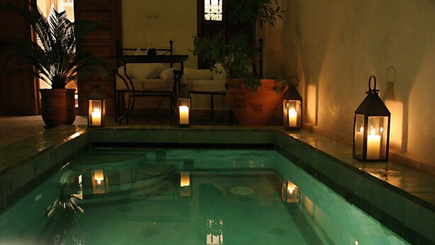 A swimming pool with a tiled edge, surrounded by potted plants and illuminated by wall-mounted lanterns. - Riad Couleurs du Sud (Marrakech)
