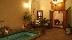 A pool area with a wooden deck, potted plants, and a red bed in the background. - Riad Couleurs du Sud (Marrakech)