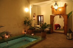 Un área de piscina con una terraza de madera, plantas en macetas y una cama roja al fondo.