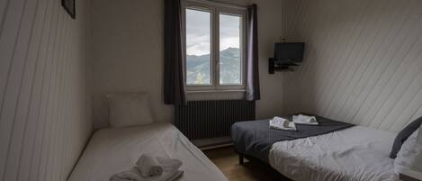 Una habitación pequeña y luminosa con dos camas, una ventana con cortinas, un televisor montado en la pared y un radiador.