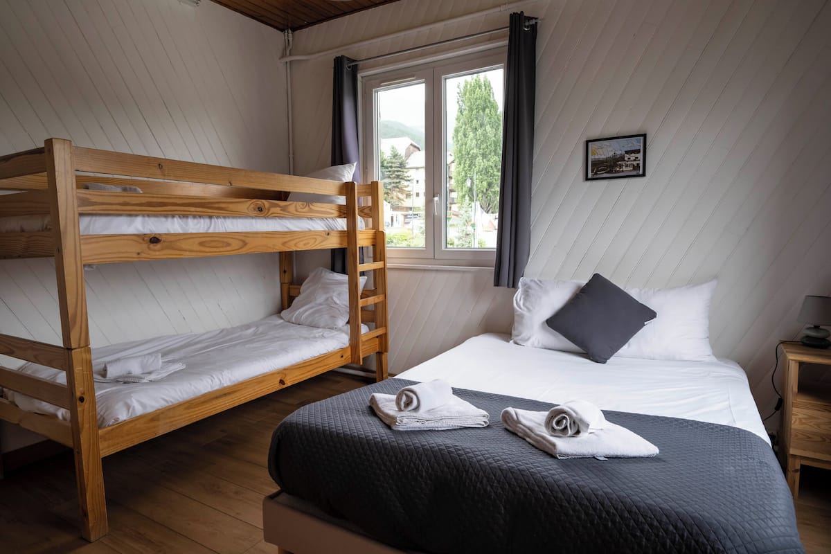 A room with a bunk bed, a single bed, a window with curtains, and a framed picture on the wall.
