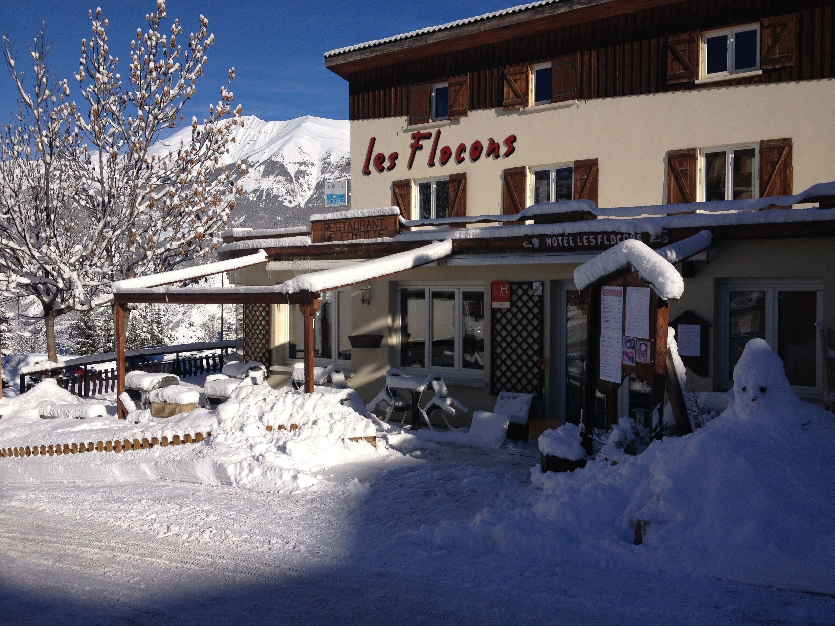 A hotel named "les Flocons" with a covered outdoor seating area and a mountain backdrop.