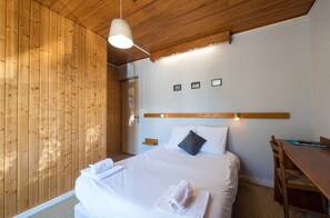 Ein ordentlich bezogenes Bett mit weißen Leinentüchern und einem gefalteten Handtuch, ein Nachttisch aus Holz, ein Holztisch und ein Wandregal mit gerahmten Bildern.