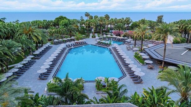 A large rectangular pool surrounded by lounge chairs and umbrellas, with palm trees and a view of the ocean. - Miramare Beach Hotel (Manavgat)