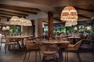 Un restaurante con mesas de madera y sillas de mimbre, grandes lámparas colgantes tejidas y un pilar de piedra.
