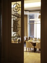 A hotel room with a traditional Chinese-style door, a dining area with patterned chairs and a table set for two, and a view of the hotel's interior through large windows.