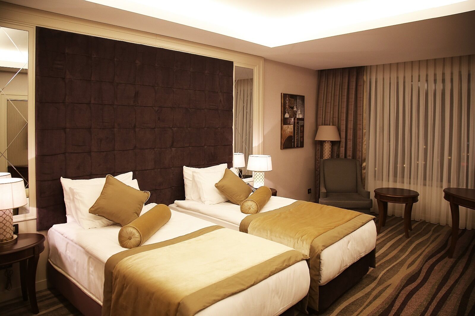 A hotel room with two beds, a large dark headboard, a chair, a small table, and a lamp.