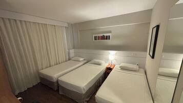 Habitación de hotel con dos camas individuales, una mesita de noche con lámpara, una pequeña mesa y una repisa con libros.