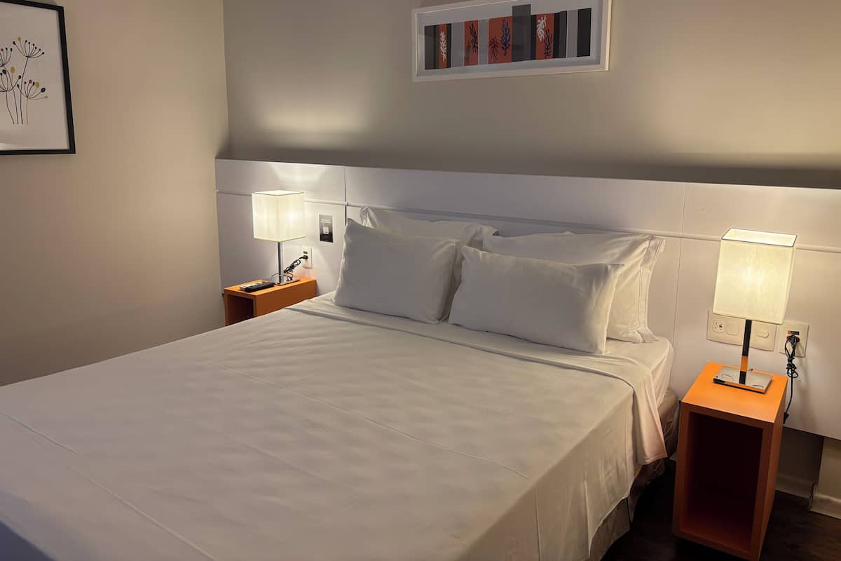 Una cama bien tendida con sábanas blancas, dos mesitas de noche con lámparas y una obra enmarcada en la pared.