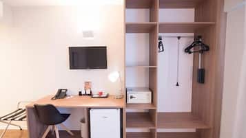 A compact office space with a desk, chair, and a small refrigerator. There is a television mounted on the wall and a shelving unit with hanging items.