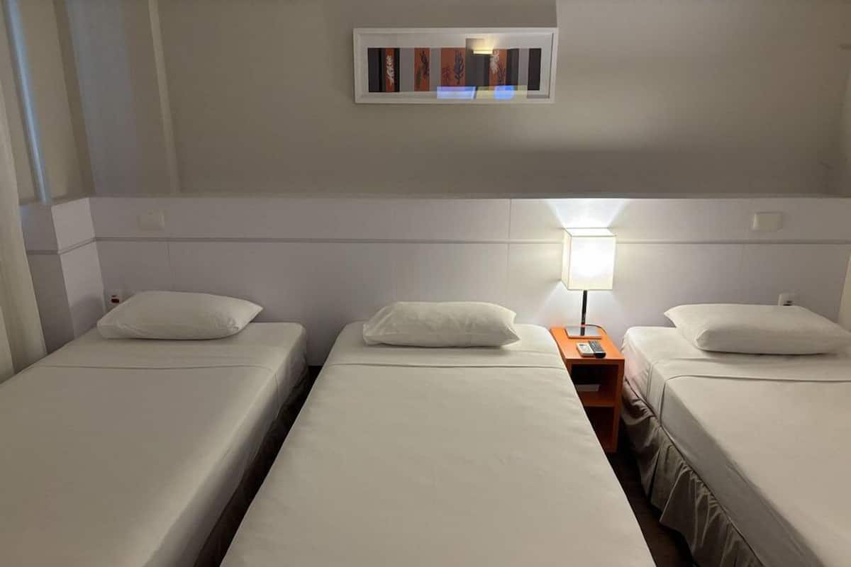 Una habitación de hotel con tres camas individuales, una mesita de noche con lámpara y una obra de arte enmarcada sobre las camas.