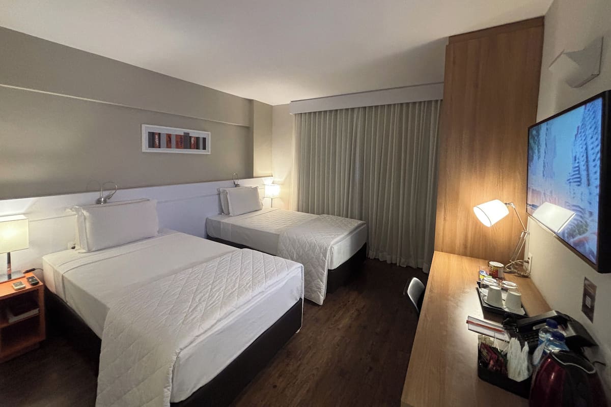 Habitación de hotel con dos camas, un escritorio con computadora, televisor y ventana con cortinas.