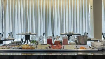 A buffet setup with various food items and beverages, a seating area with tables and chairs, and a large window with vertical blinds.