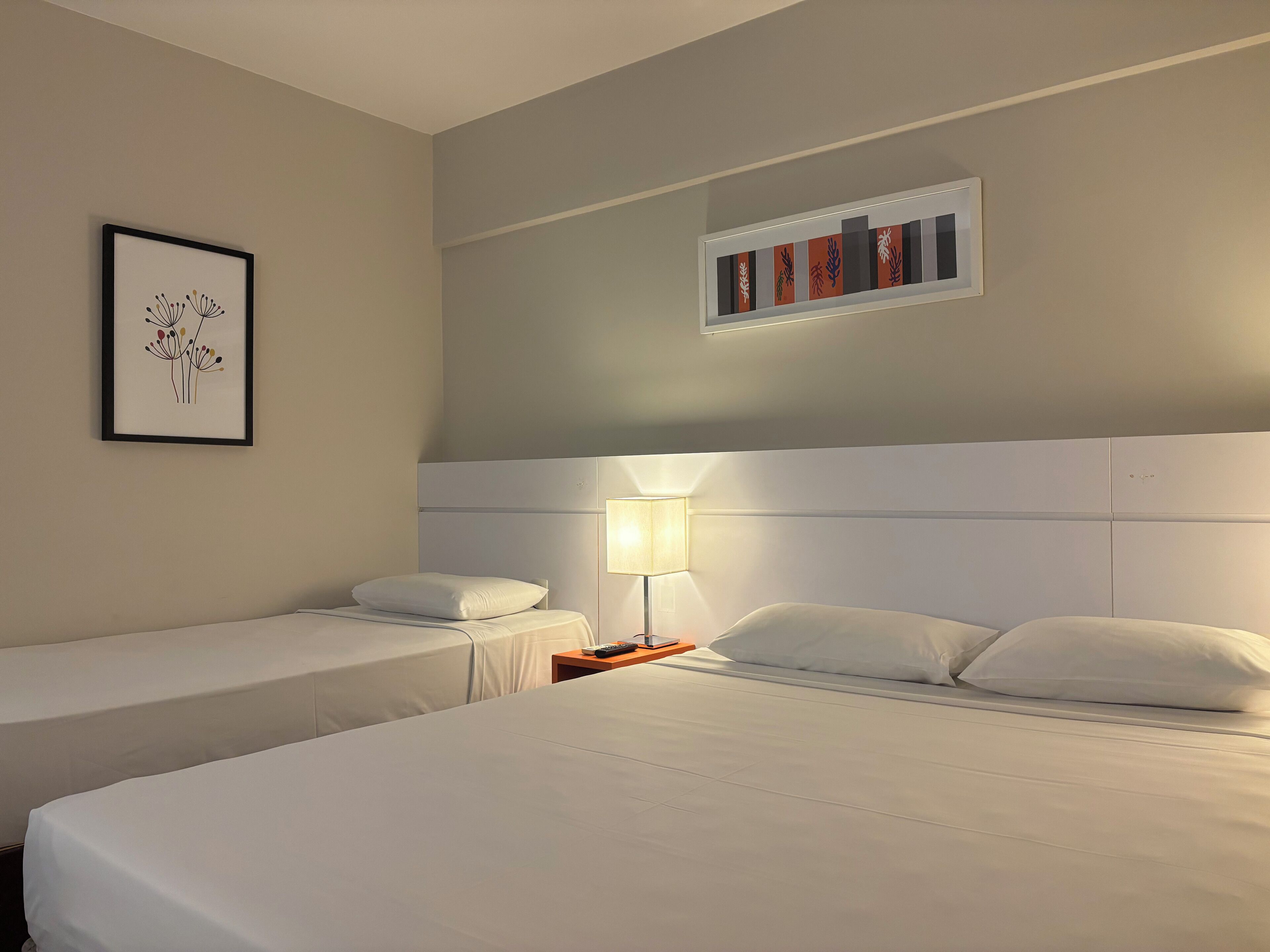 A hotel room with two single beds, a nightstand with a lamp, a framed picture on the wall, and a bookshelf with books.