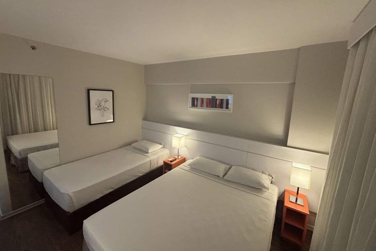 Habitación de hotel con dos camas individuales, una cama doble, una mesita de noche con lámpara, un cuadro en la pared y un estante pequeño con libros.