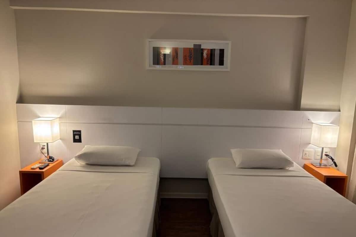 Habitación de hotel con dos camas individuales, cada una con una mesita de noche y una lámpara. Encima de las camas hay un pequeño estante decorado con objetos.