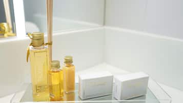 A bathroom with a glass shelf displaying toiletries, including a bottle of Trousseau body wash and two boxes of Trousseau body soap.