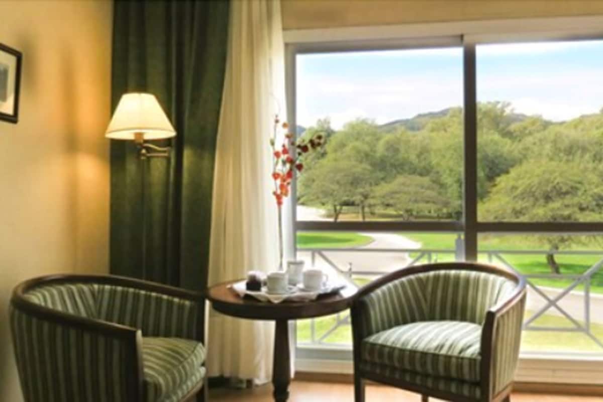 A room with two striped chairs, a table with a tray of items, a lamp, and a view of a green landscape through large windows.