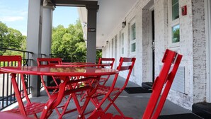 A covered outdoor seating area with red metal chairs and a glass table, white brick walls, and a door with a sign.