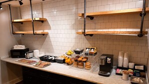 A kitchen area with a marble countertop, black cabinets, and a brick wall. There are shelves with various food items, a coffee machine, and a microwave.