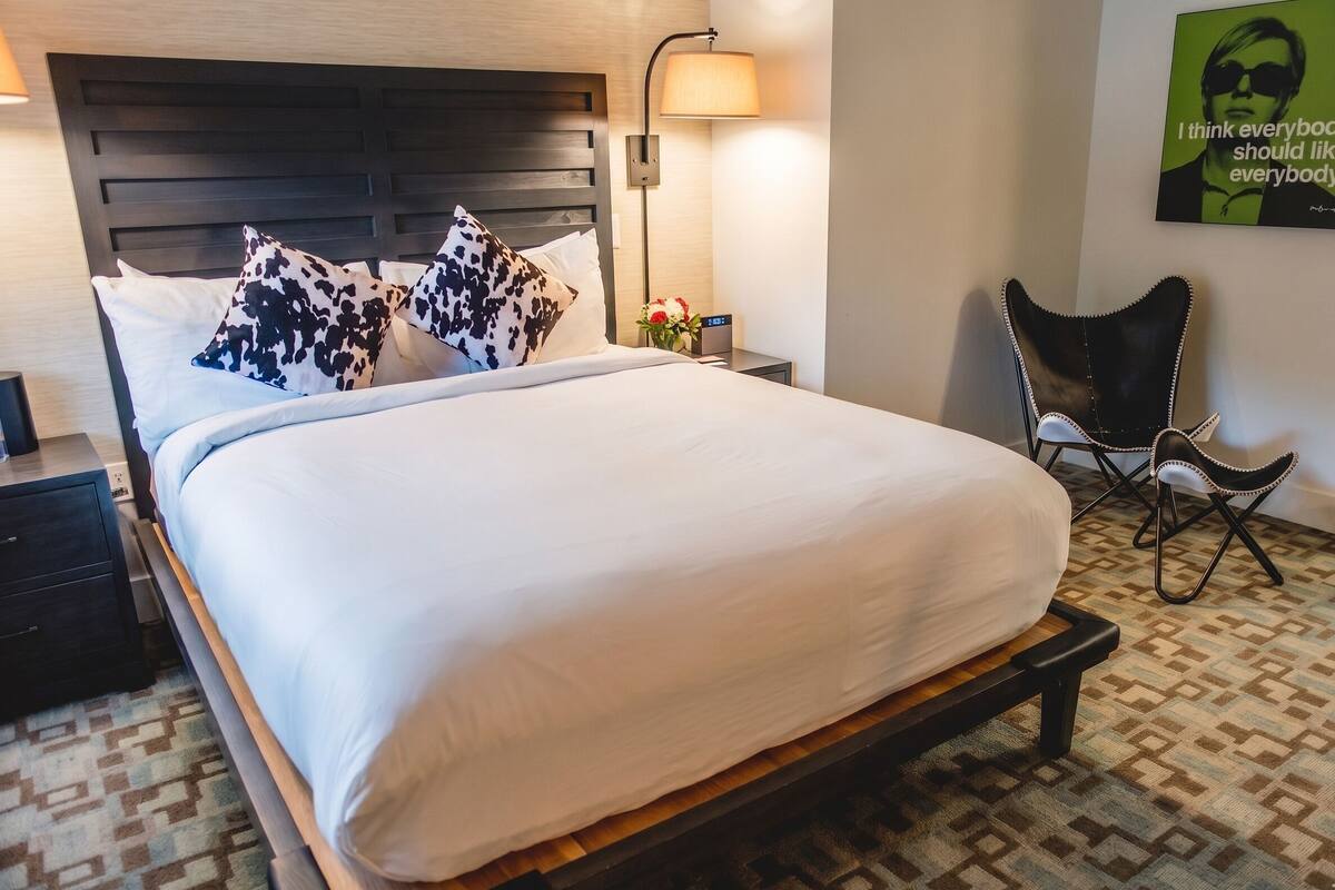 A neatly made bed with a headboard, a bedside lamp, a chair, and a wall clock.