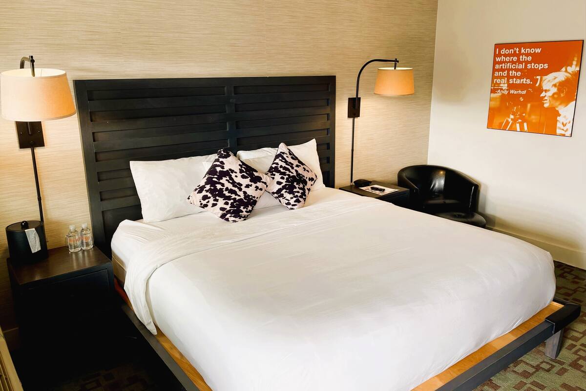 A hotel room with a bed, two bedside lamps, a nightstand with a phone and a lamp, and a poster on the wall.
