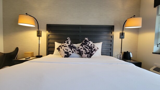 A hotel room with a large bed, two bedside lamps, a wooden headboard, and a nightstand with a phone and a remote control.