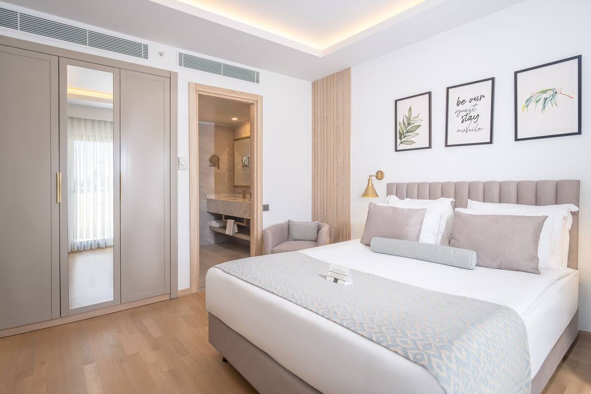 A modern hotel room with a large bed, a wardrobe, a bathroom, and a bedside table with a lamp.