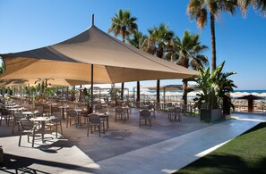 An outdoor café area with tables and chairs under a large canopy, surrounded by palm trees and a view of the beach. - Miramare Queen Hotel - All Inclusive (Manavgat)