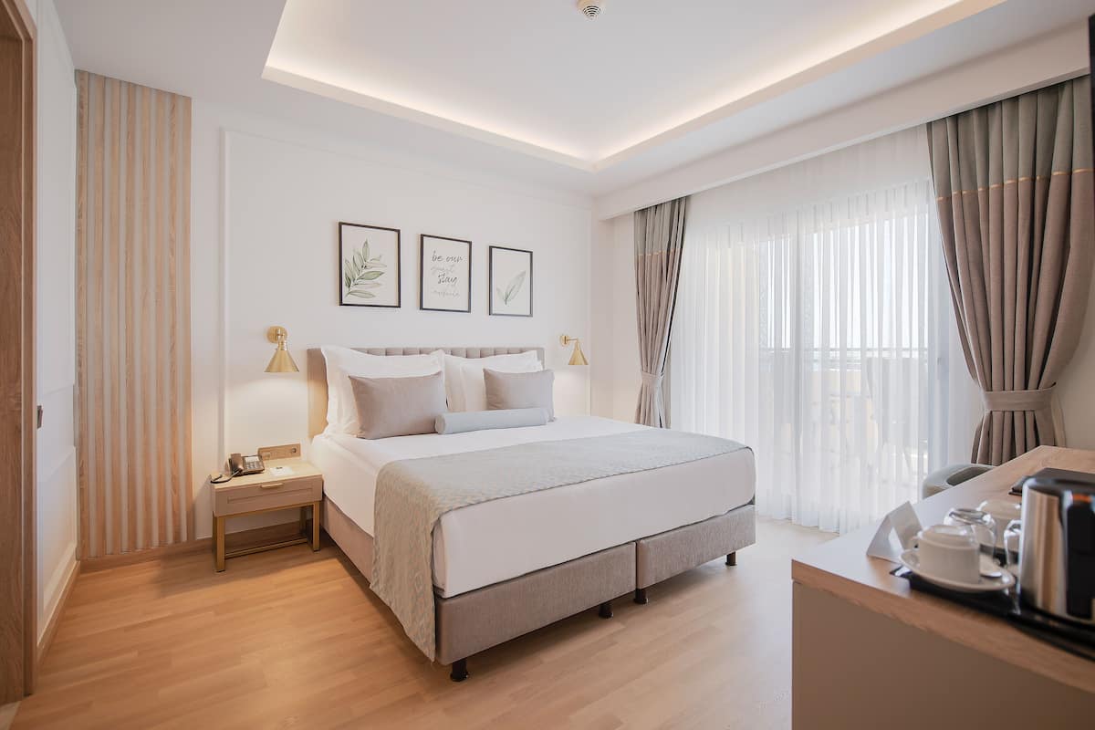 A modern hotel room with a large bed, bedside tables, a desk with a kettle, and curtains.