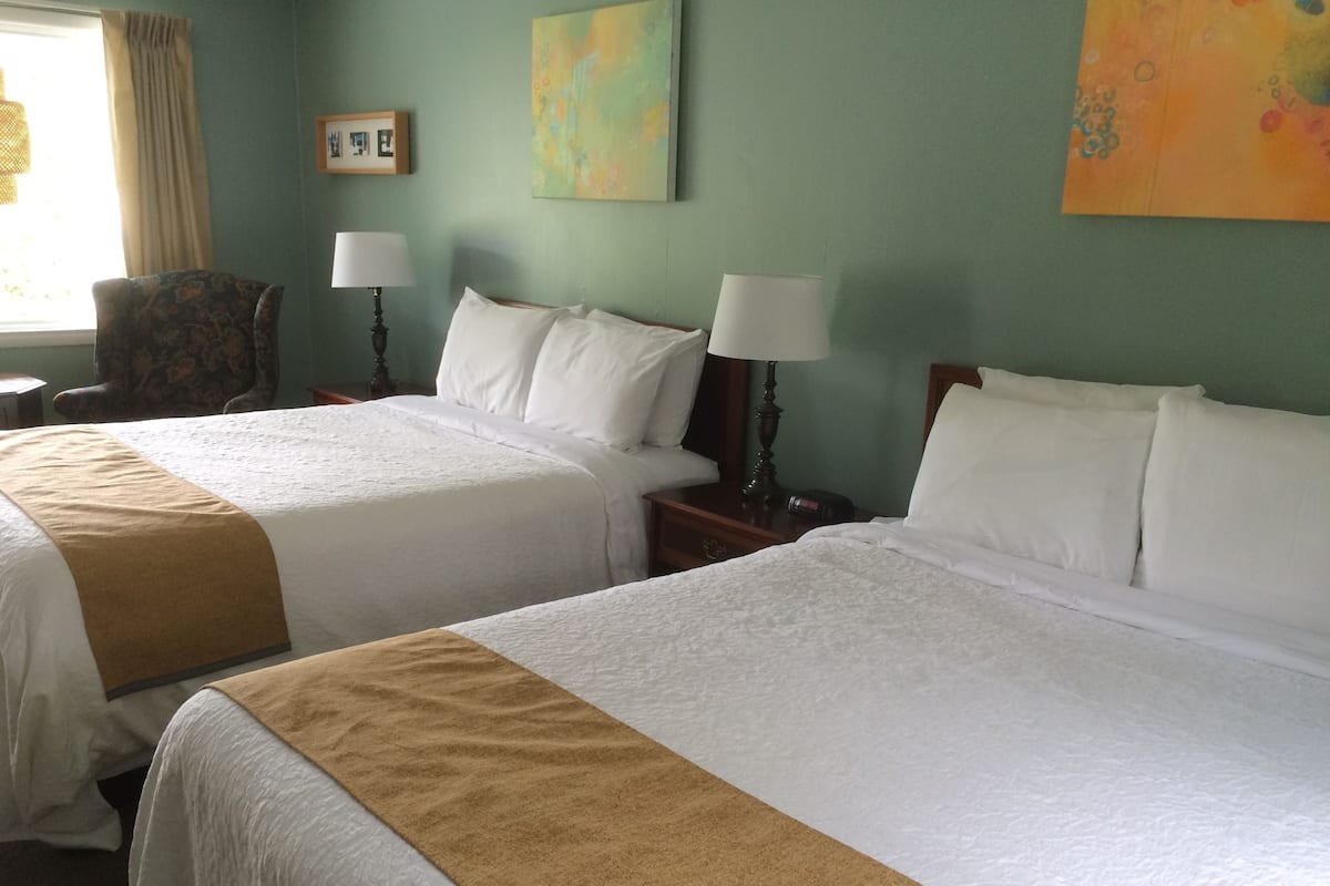 A hotel room with two beds, each with white linens and a brown throw blanket. There are two bedside tables with lamps, a chair, and a painting on the wall.