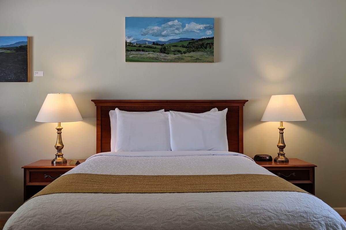 A neatly made bed with white linens, flanked by two bedside tables with lamps, and a landscape painting on the wall above.