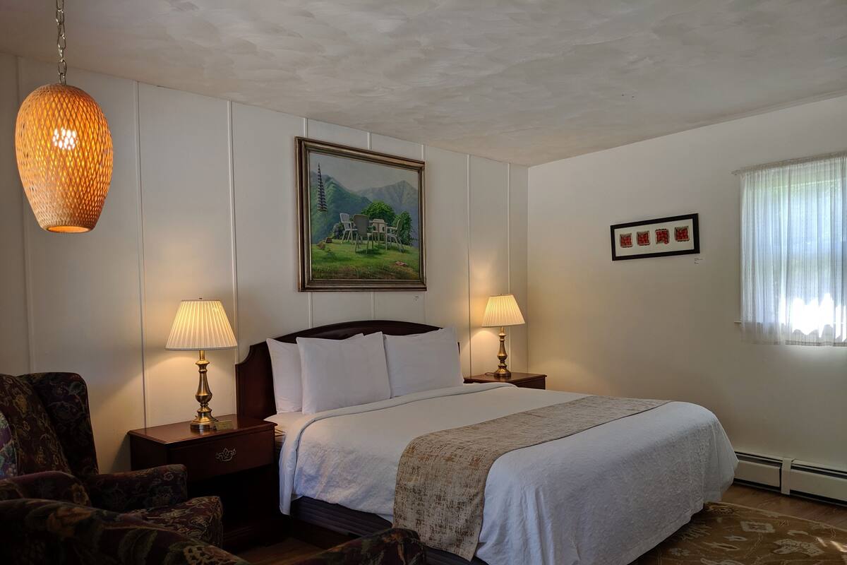 A hotel room with a large bed, two bedside lamps, a framed landscape painting, a patterned rug, and a hanging light fixture.