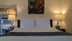 A hotel room with a bed, two bedside lamps, a painting on the wall, and an air conditioning unit. - Harwood Hill Motel (Bennington)