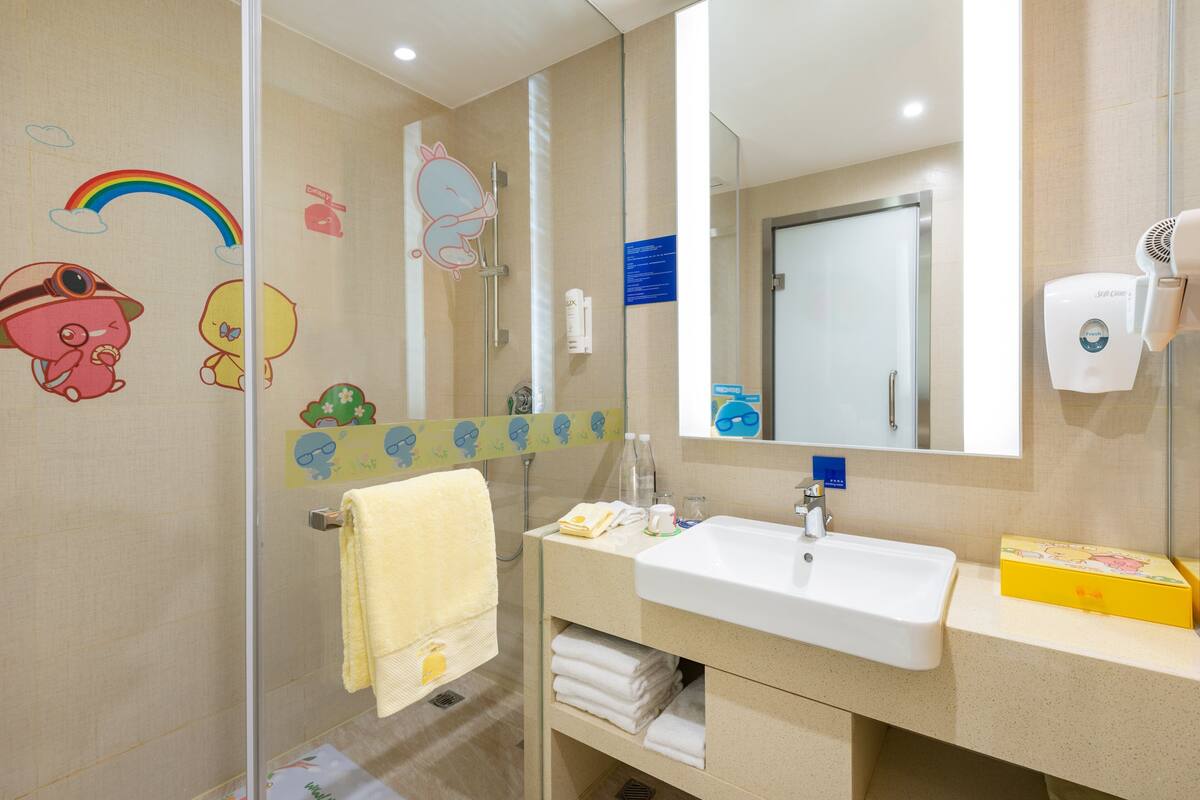 A bathroom with a white sink, mirror, and glass shower enclosure. There are cartoon stickers on the walls and a yellow towel rack.