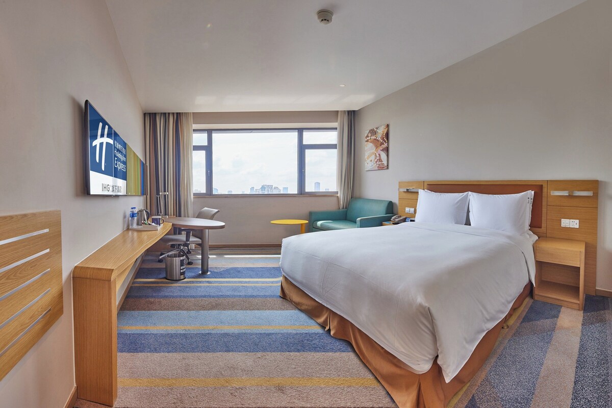 A hotel room with a bed, a desk, a chair, a small table, and a window with a city view.