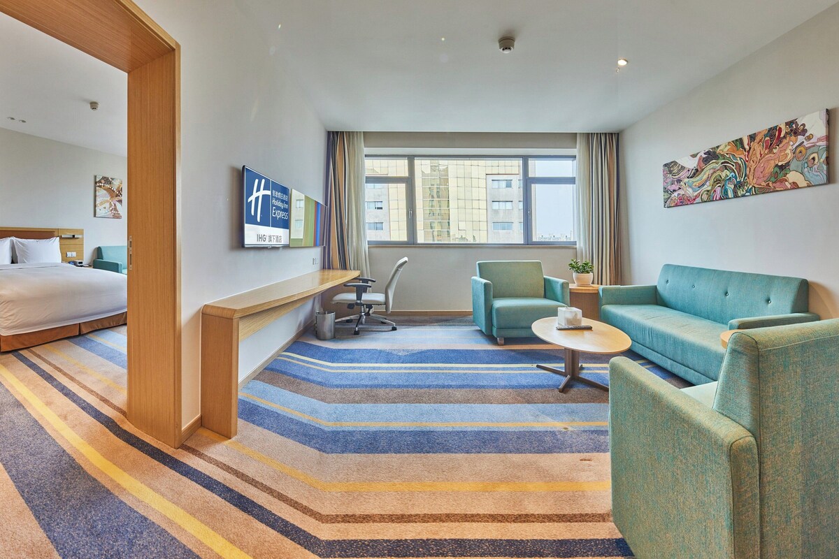 A hotel room with a bed, a desk, a chair, a sofa, a small table, and a window with a view of buildings.