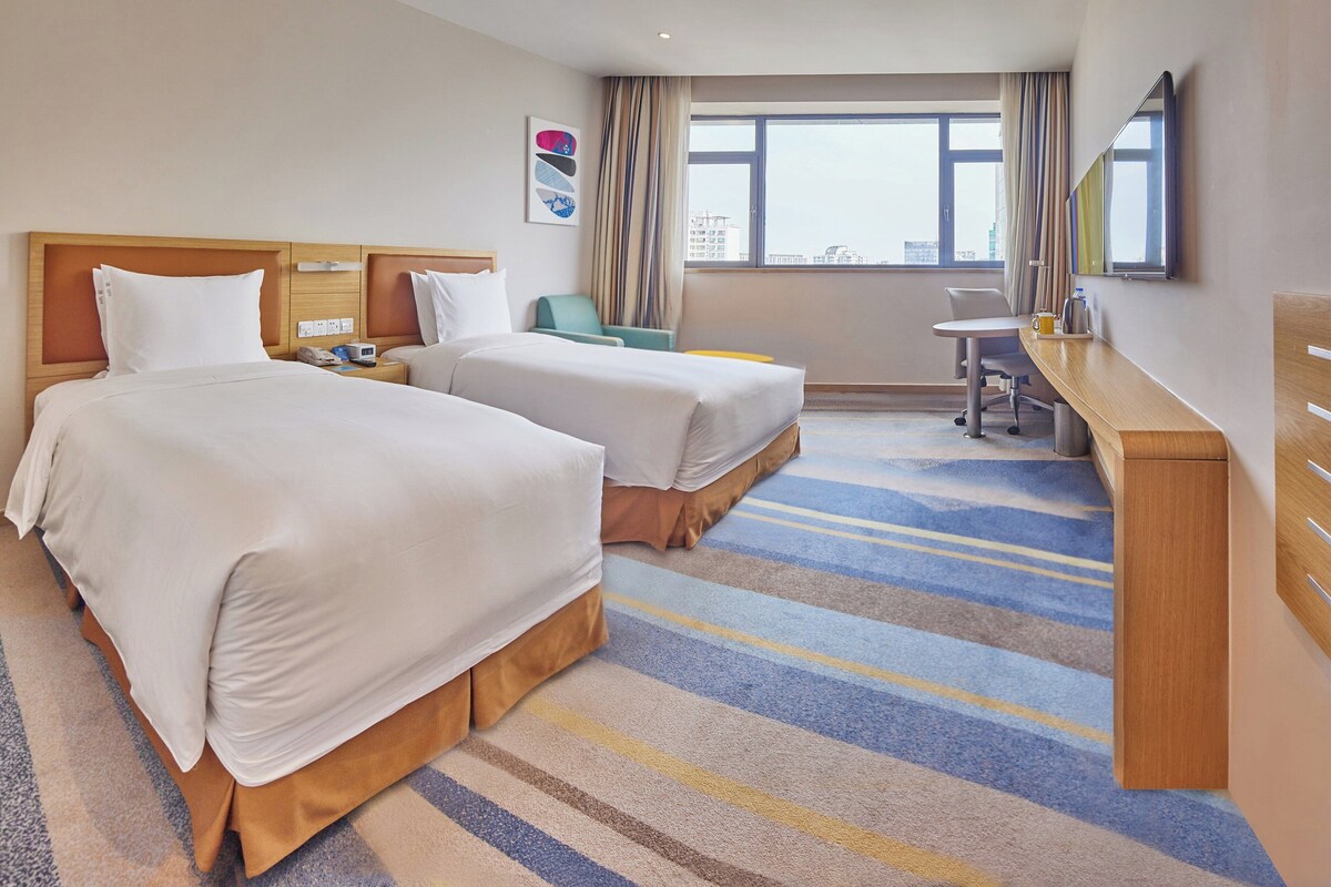 A hotel room with two beds, a desk, a chair, and a large window offering a city view.