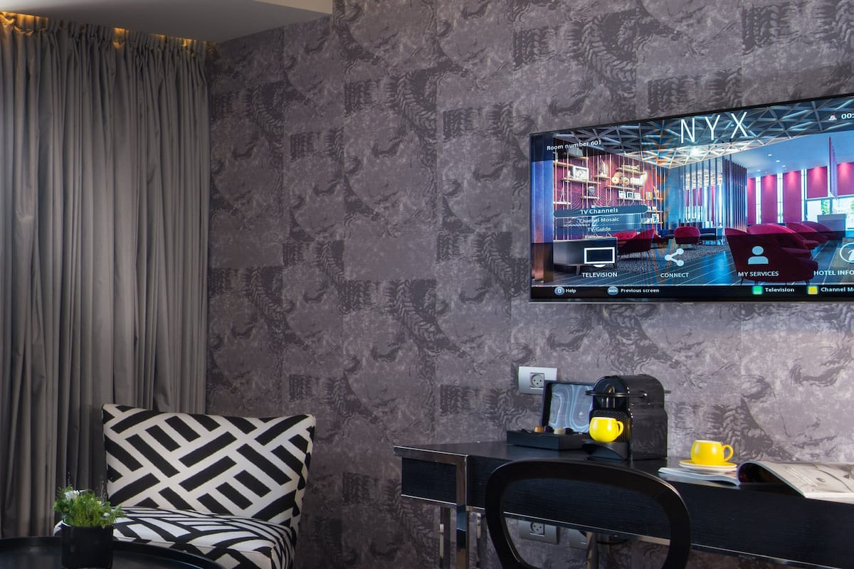 A modern hotel room with a large flat-screen TV displaying a room layout, a desk with a coffee maker, a chair, and a decorative pillow.