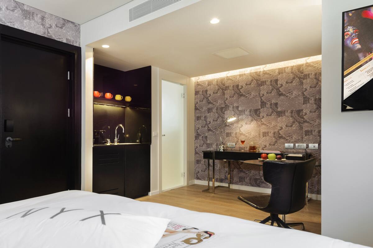 A modern hotel room with a bed, a desk with a chair, a small kitchenette, and a bathroom.